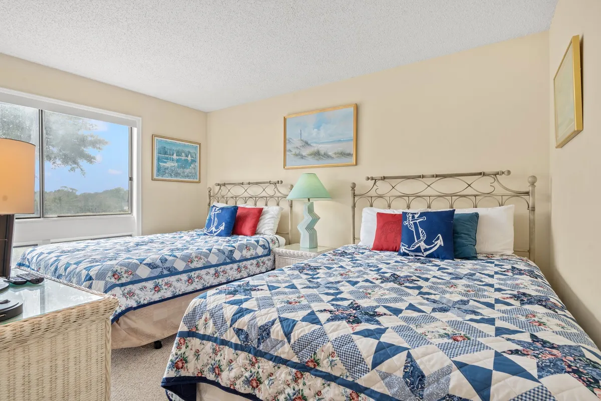 A comfortable bedroom setup with two beds featuring patterned quilts, a colorful lamp between them, and framed artwork on the wall. The room is designed for both individual comfort and shared experiences, accommodating families or groups.