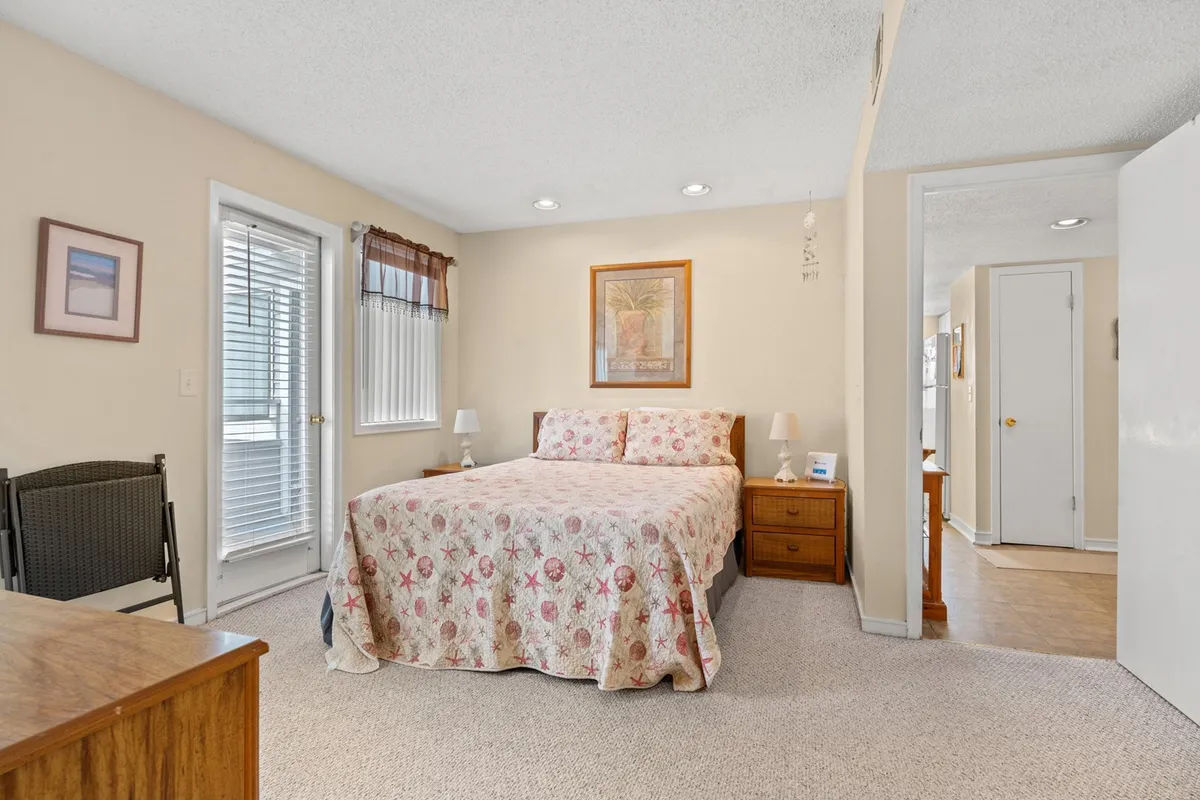 A cozy bedroom featuring a queen bed adorned with a floral quilt, complemented by tasteful wall art and a window letting in natural daylight. A nightstand with a lamp sits beside the bed, creating an inviting space for rest or quiet reading.