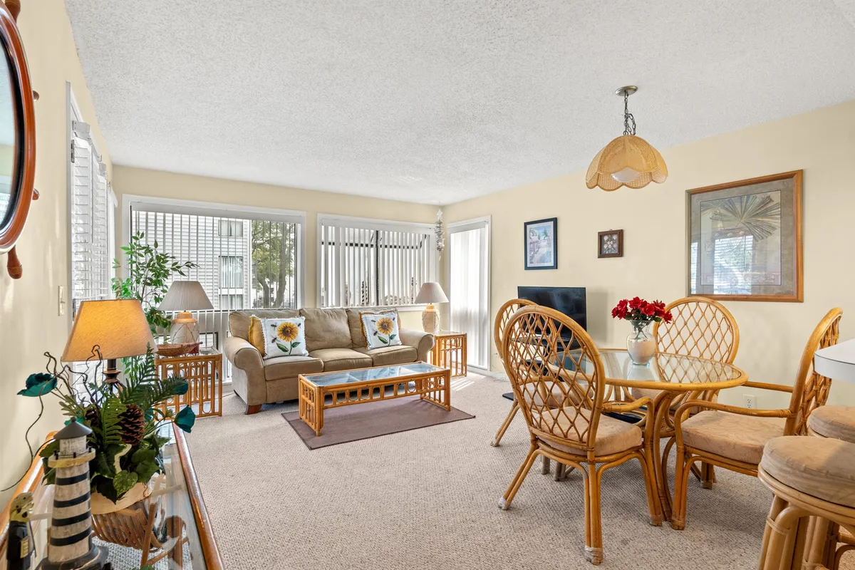 The image showcases a bright and cozy open living and dining area, featuring a comfortable sofa adorned with colorful decorative cushions, a wooden dining table with rattan chairs, a softly glowing lamp, and stylish framed wall art. The inviting setup hints at shared family meals, relaxed mornings with coffee, and memorable gatherings with friends.
