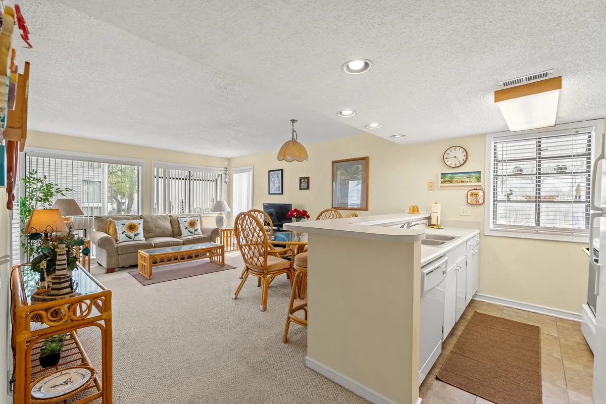 This image features a cozy living room with a sofa adorned with decorative pillows, a dining table with chairs set for gathering, and a kitchen bar area visible in the background. Warm lighting from an overhead fixture enhances the inviting atmosphere, while the open layout showcases how the living and dining spaces are connected.
