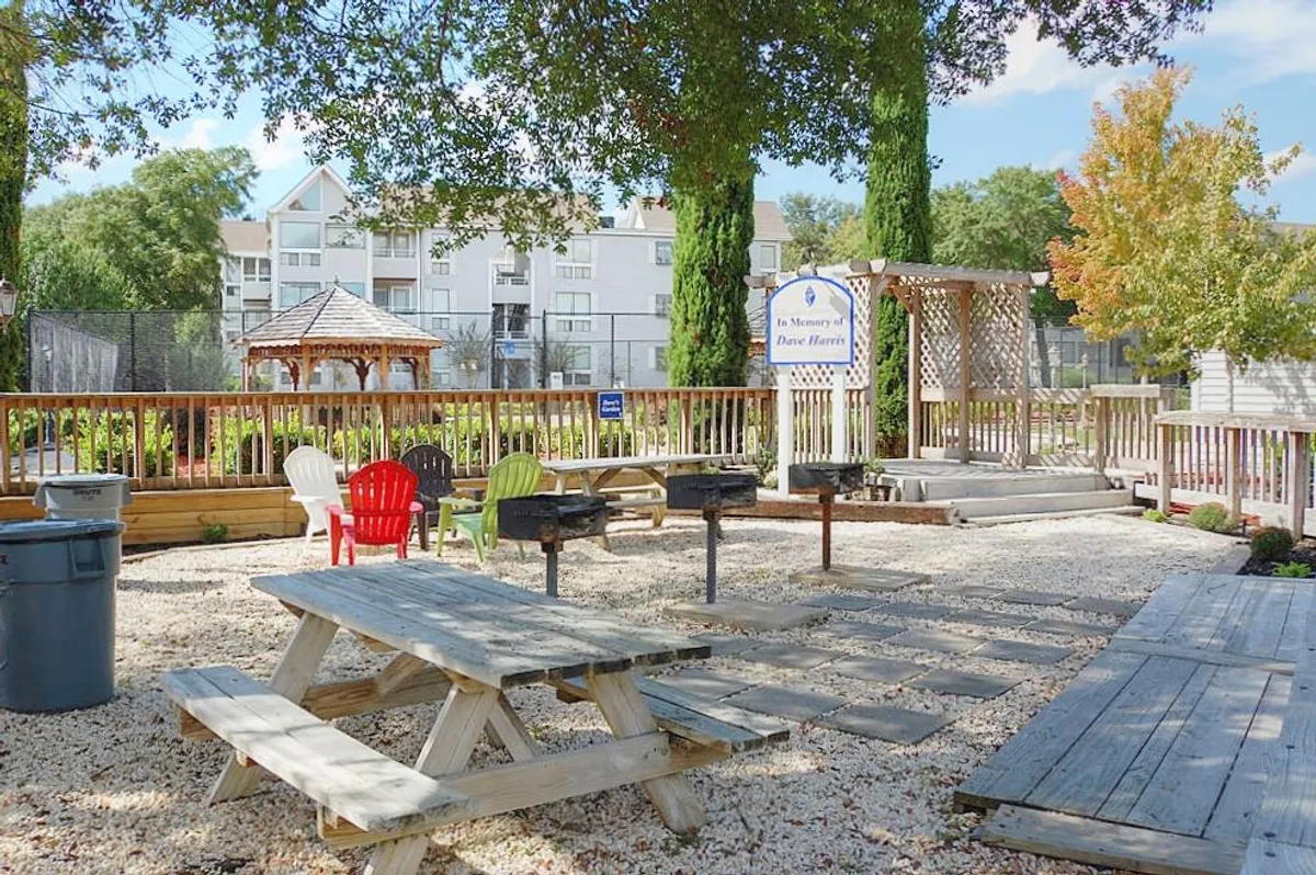 A well-maintained outdoor grilling and picnic area featuring multiple grill stations, colorful chairs, picnic tables under a pergola, and lush landscaping. The space looks perfect for social gatherings, barbecues, and relaxed meals with friends or family.