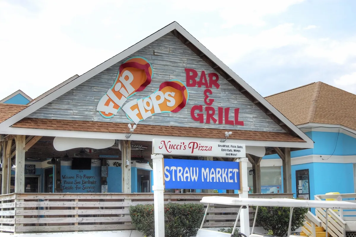 A casual, inviting strip featuring signage for Flip Flops Bar & Grill, Nucci's Pizza, and the Straw Market—popular local spots for dining, shopping, and socializing. The mood is laid-back and friendly, perfect for unwinding after a day at the beach.