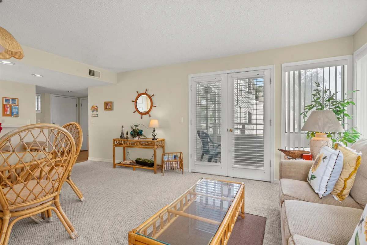 A welcoming living space furnished with woven chairs, a glass coffee table, a decorative mirror, and indoor plants. Large sliding doors let in abundant sunlight, suggesting a seamless connection to the private balcony beyond. The cozy arrangement invites relaxation and socializing.