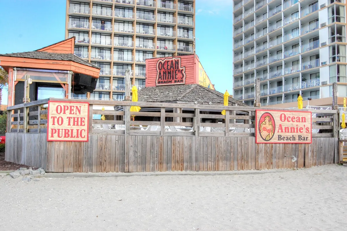 The open, sunny patio of Ocean Annie’s Beach Bar with cheerful yellow umbrellas and friendly signage. The scene is alive with the promise of social gatherings, group relaxation, and easy access to fun outings just steps from Arcadian Dunes.