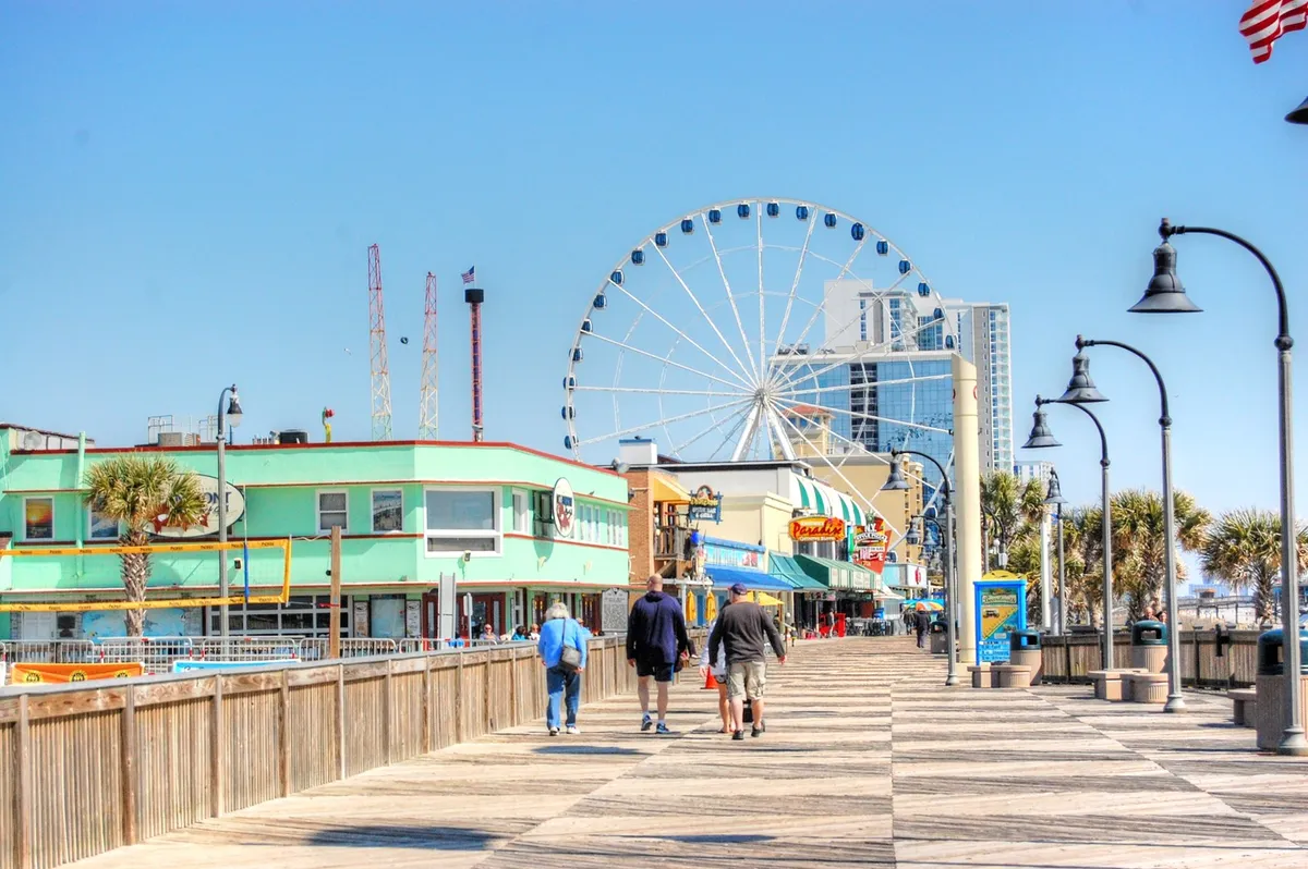 A vibrant Myrtle Beach boardwalk scene featuring a towering ferris wheel, colorful buildings, palm trees, and crowds strolling along the walkway. The scene is energetic and joyful, representing popular attractions like the SkyWheel, local shops, and entertainment options mentioned in the section.