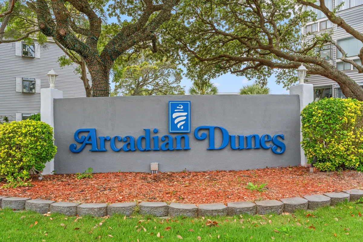 This image features the Arcadian Dunes sign framed by lush greenery and a neat stone border, immediately signaling the entrance and welcoming atmosphere of the property. The scene feels inviting and hints at a tranquil, well-maintained environment, perfect for guests seeking an extended stay.