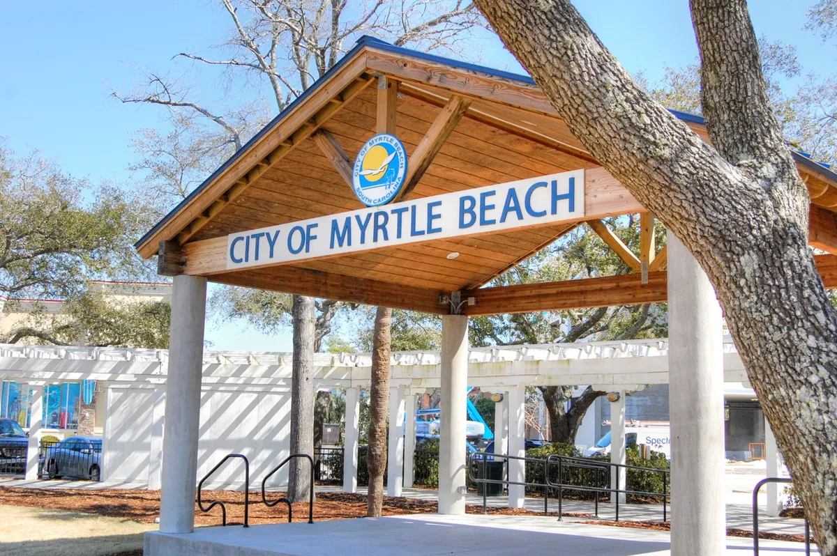 The image showcases the 'CITY OF MYRTLE BEACH' signage set on a wooden pavilion structure, surrounded by green trees. It hints at a lively community space where local events and gatherings occur, illustrating the area's rich community and social opportunities.