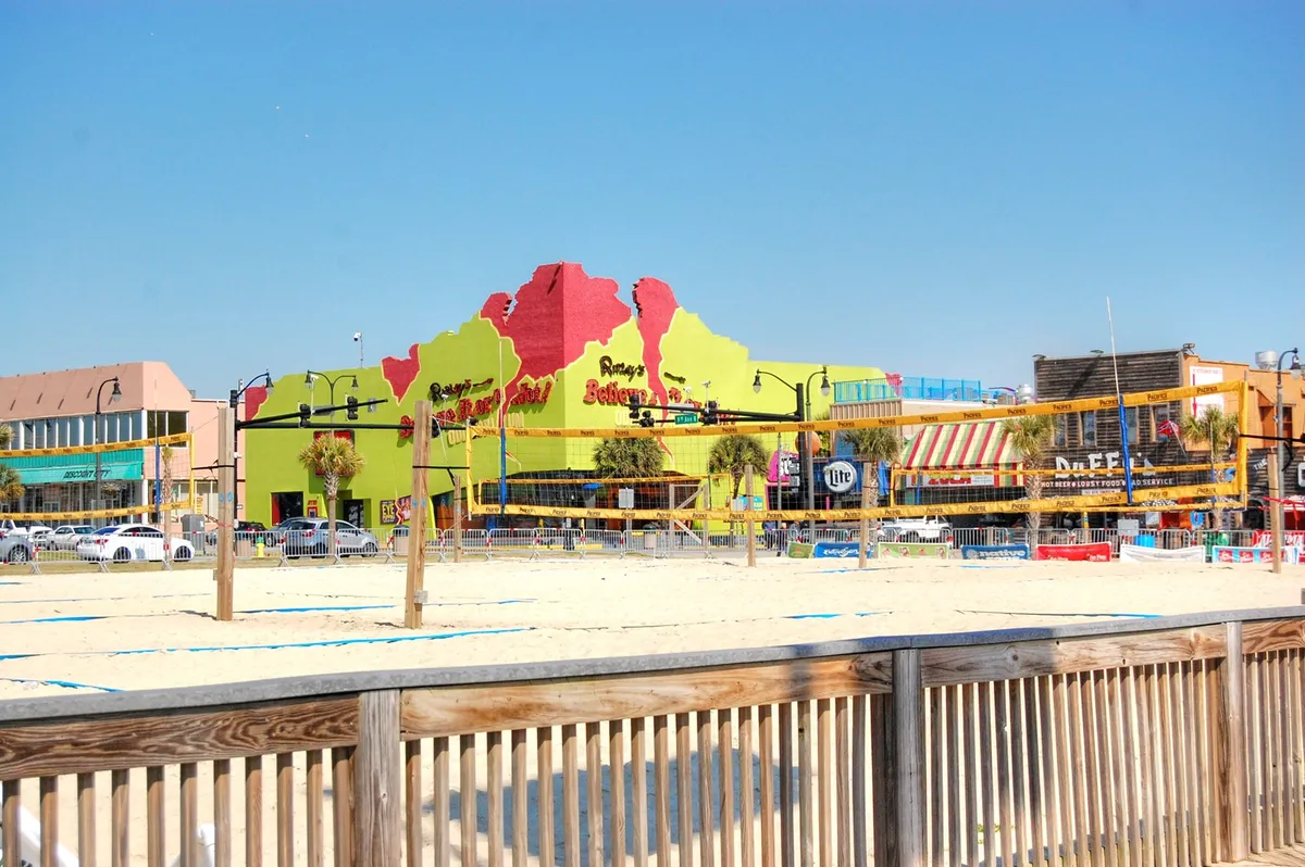 A vibrant exterior scene with a colorful building facade, adjacent beach volleyball court, and palm trees, evoking a festive atmosphere just steps from the sand. The lively setting hints at beachside fun, community events, and energetic nightlife.