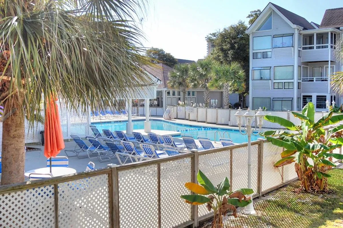 A sparkling swimming pool is surrounded by palm trees, lounge chairs, and an orange umbrella. The scene is sunny and inviting, with people relaxing poolside and enjoying the vibrant, resort-like atmosphere.