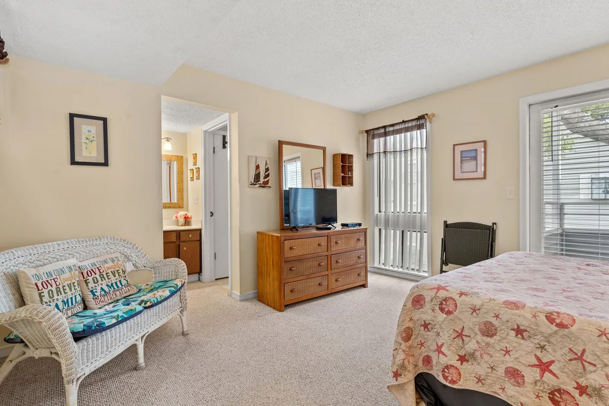 A bright, welcoming living space featuring a bed with a starfish-patterned blanket, wicker loveseat adorned with pillows, a TV on a wooden dresser, and a sunlit window. The room invites relaxation, reading, or socializing, echoing the comfort of a home away from home.