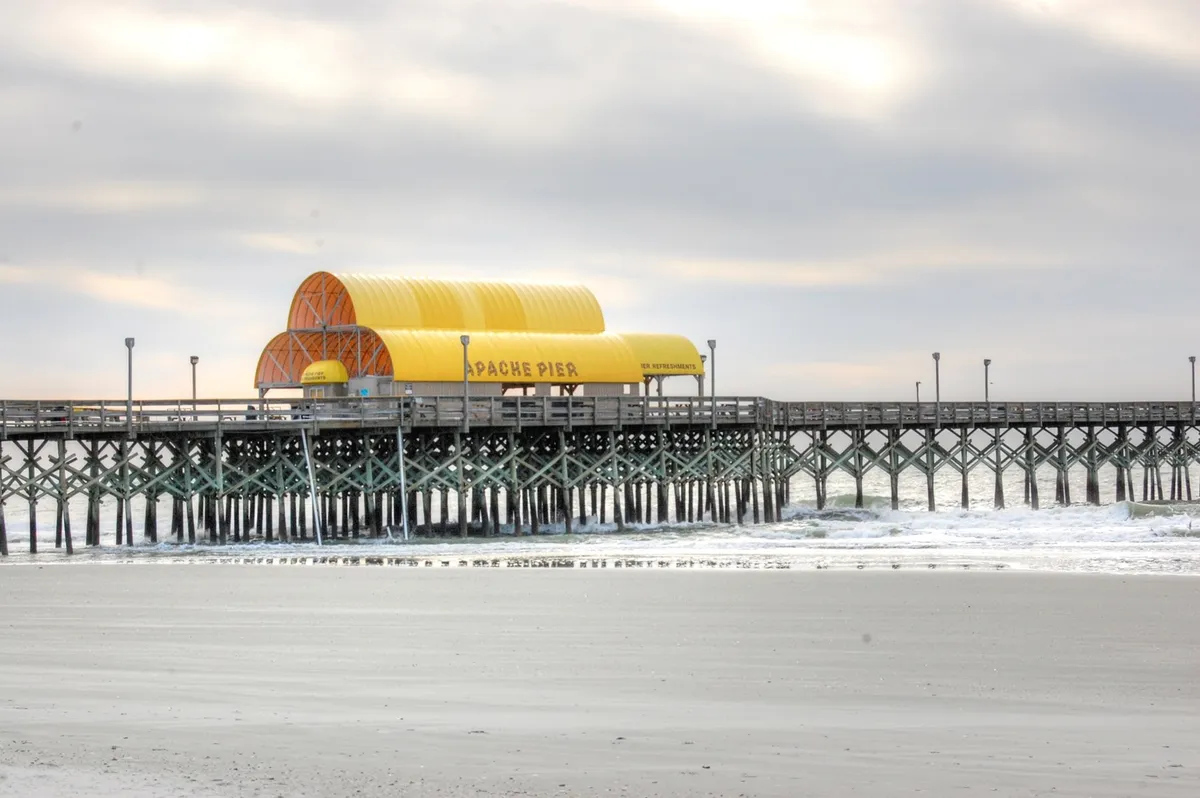 This image captures the iconic Apache Pier stretching out over the beach and ocean under a cloudy sky. The pier is a well-known landmark in Myrtle Beach, perfect for fishing, scenic walks, and capturing the essence of a beachside vacation. The view includes sandy shores and gentle ocean waves, emphasizing proximity to the water and outdoor fun.