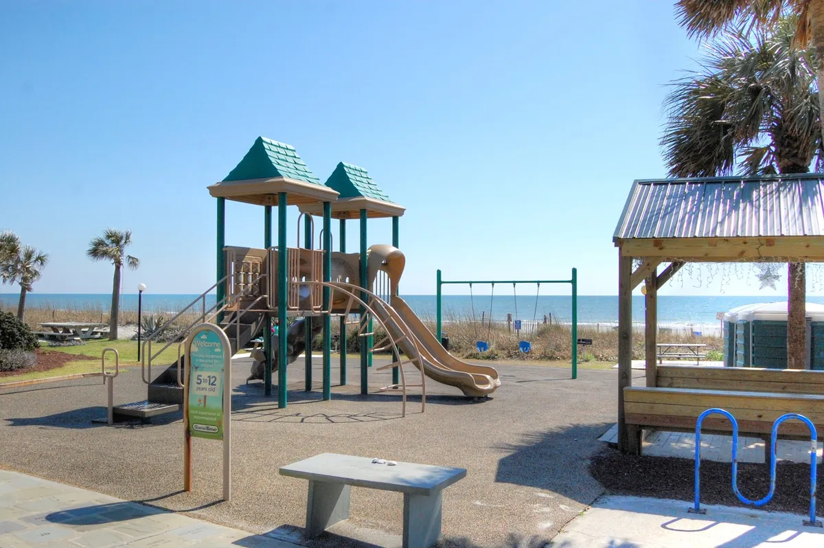 The scene showcases a beachfront playground with bright equipment, swings, benches, and a view of the ocean in the background. Children play while families gather for picnics and enjoy the carefree beach atmosphere. This space illustrates the family-friendly environment and the fun awaiting young guests.